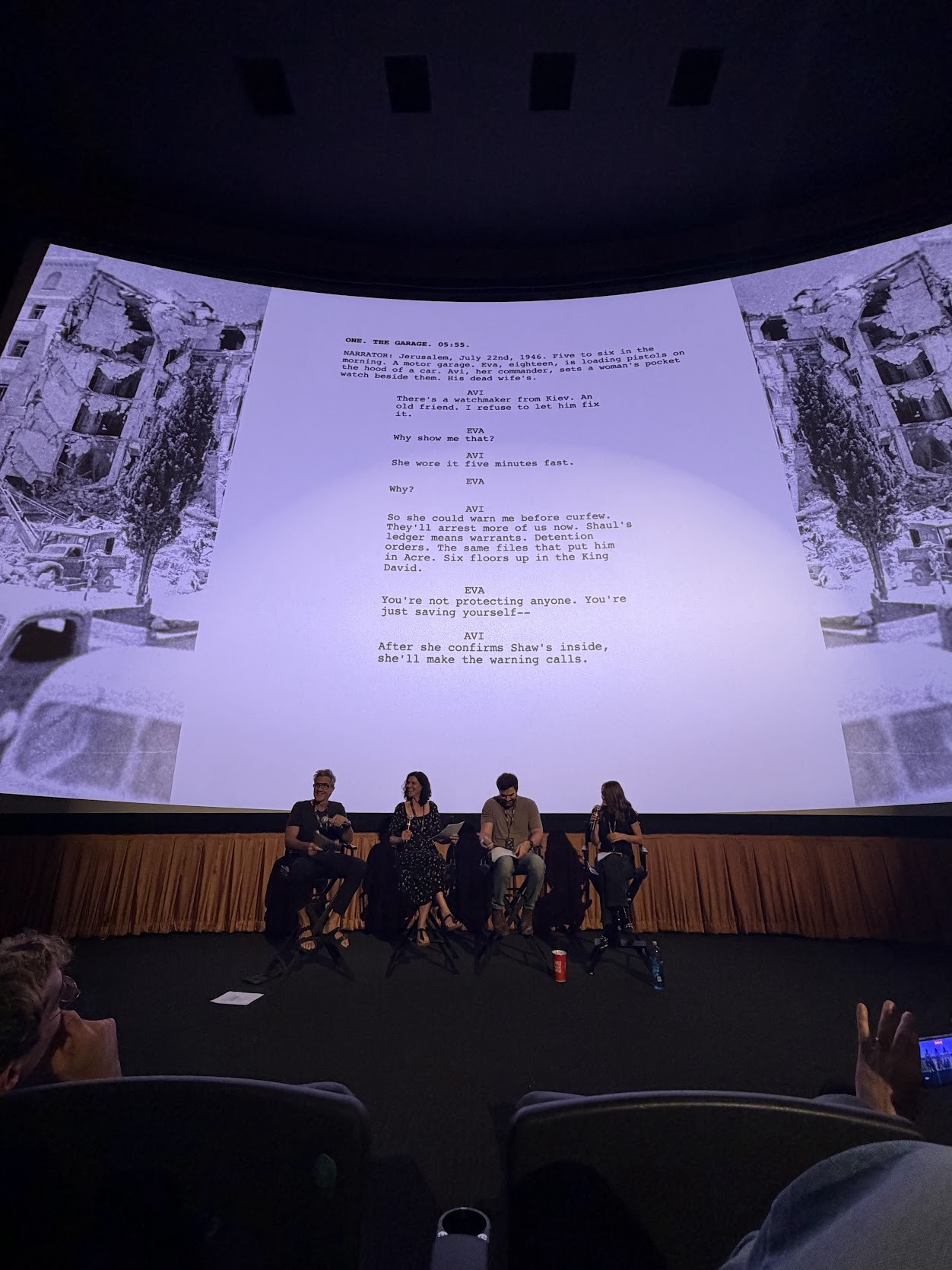 Four actors seated on stage, seen from the front row, with the opening scene of the 12:37 pilot projected on the screen behind them.
