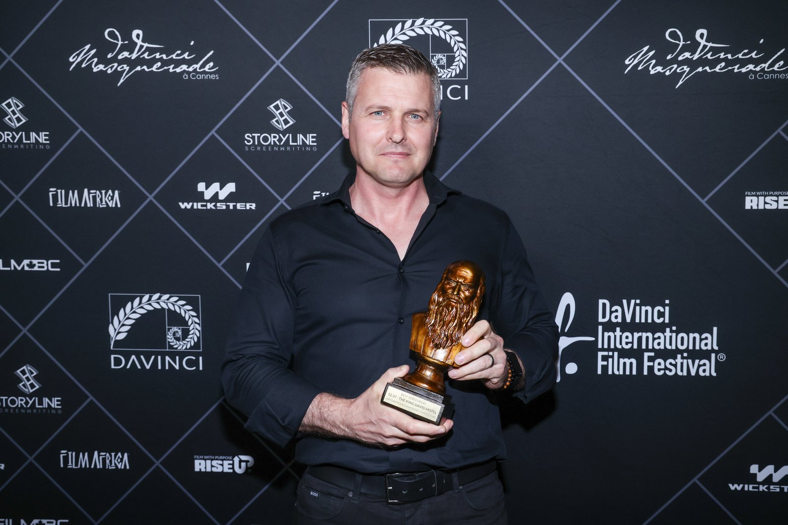 Michael John Fedun holding the Leo Award, a bronze bust, in front of the DaVinci International Film Festival backdrop. The plaque reads: Best Screenplay, 12:37 &ndash; The King David Hotel, 2026 DaVinci International Film Festival.